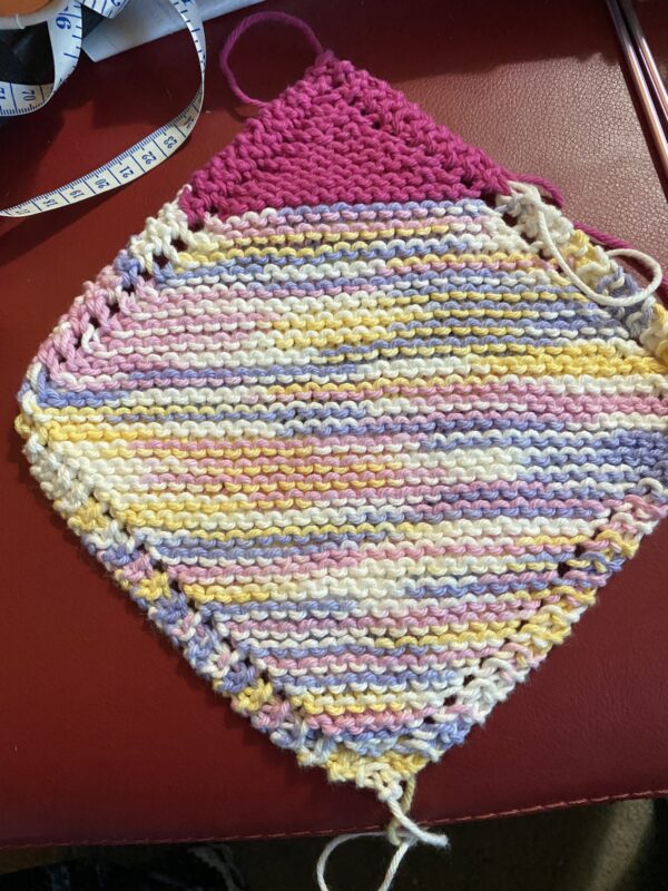 A square garter stitch washcloth knit in variegated white, yellow, pink and blue with one corner in deep pink.